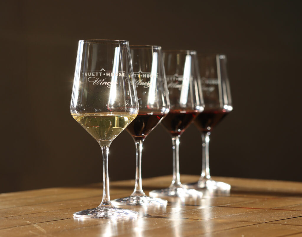 Four engraved Truett-Hurst Winery glasses in a row on a wood table, one of white wine and three of red, in warm side light